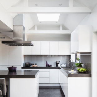 Photo of a medium sized contemporary l-shaped kitchen/diner in Sussex with white cabinets, engineered stone countertops, black appliances, a breakfast bar and grey worktops.