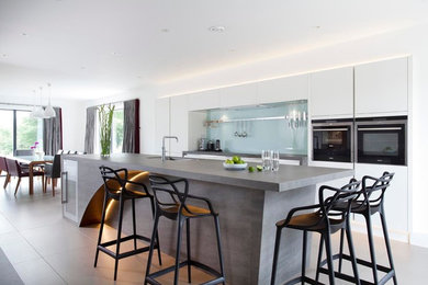 This is an example of an expansive contemporary u-shaped open plan kitchen in Other with an integrated sink, flat-panel cabinets, white cabinets, engineered stone countertops, green splashback, glass sheet splashback, stainless steel appliances, ceramic flooring and an island.