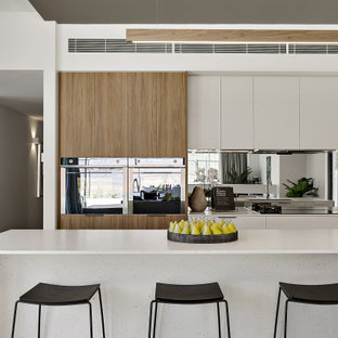 Design ideas for a mid-sized contemporary single-wall eat-in kitchen in Canberra - Queanbeyan with an undermount sink, flat-panel cabinets, white cabinets, quartz benchtops, metallic splashback, mirror splashback, black appliances, concrete floors, with island, grey floor and white benchtop.
