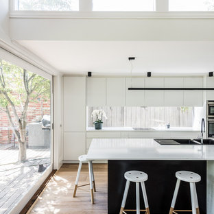 Inspiration for a modern galley kitchen in Melbourne with a drop-in sink, flat-panel cabinets, white cabinets, window splashback, stainless steel appliances, medium hardwood floors, with island, brown floor and white benchtop.