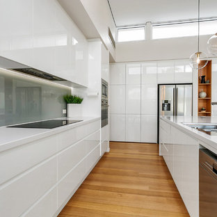Contemporary u-shaped kitchen in Adelaide with flat-panel cabinets, white cabinets, grey splashback, glass sheet splashback, stainless steel appliances, medium hardwood floors, with island, brown floor and white benchtop.