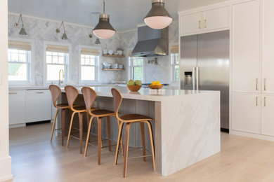 Large beach style l-shaped light wood floor and beige floor open concept kitchen photo in Boston with a drop-in sink, recessed-panel cabinets, white cabinets, quartz countertops, marble backsplash, stainless steel appliances, an island, white countertops and gray backsplash