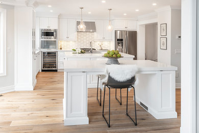 Mid-sized trendy l-shaped medium tone wood floor and brown floor kitchen photo in Toronto with an undermount sink, recessed-panel cabinets, white cabinets, stainless steel appliances, two islands, quartzite countertops, white backsplash, marble backsplash and white countertops