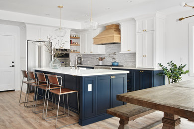 Open concept kitchen - large coastal l-shaped light wood floor and brown floor open concept kitchen idea in San Francisco with shaker cabinets, gray backsplash, stainless steel appliances, an island, a farmhouse sink, white cabinets, solid surface countertops and white countertops