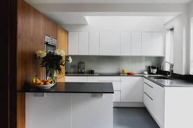 Contemporary kitchen in London with a submerged sink, flat-panel cabinets, white cabinets, grey splashback, glass sheet splashback, stainless steel appliances, a breakfast bar, grey floors and black worktops.