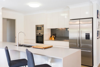 This is an example of a large modern galley open plan kitchen in Brisbane with a submerged sink, flat-panel cabinets, beige cabinets, engineered stone countertops, grey splashback, glass sheet splashback, stainless steel appliances and an island.