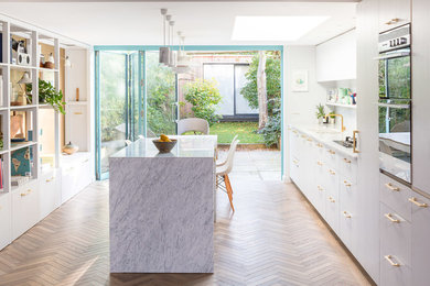 Design ideas for a contemporary single-wall kitchen/diner in London with a built-in sink, flat-panel cabinets, white cabinets, marble worktops, white splashback, marble splashback, stainless steel appliances, medium hardwood flooring, an island and brown floors.