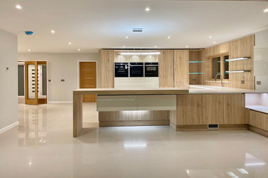 This is an example of a large contemporary u-shaped open plan kitchen in Devon with a submerged sink, flat-panel cabinets, white cabinets, composite countertops, brown splashback, wood splashback, black appliances, porcelain flooring, a breakfast bar, beige floors and white worktops.