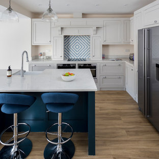 Photo of a medium sized contemporary l-shaped kitchen in London with a belfast sink, shaker cabinets, grey cabinets, multi-coloured splashback, stainless steel appliances, light hardwood flooring, an island, white worktops, ceramic splashback and brown floors.