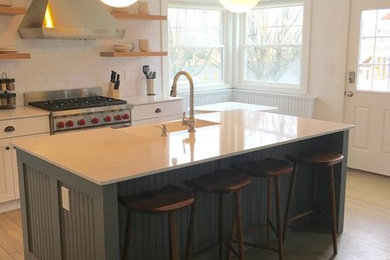 Example of a large transitional l-shaped light wood floor and brown floor open concept kitchen design in New York with quartz countertops, a farmhouse sink, white backsplash, subway tile backsplash, stainless steel appliances, an island, white countertops, shaker cabinets and white cabinets