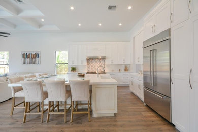 Example of a large trendy l-shaped medium tone wood floor and brown floor open concept kitchen design in Miami with a farmhouse sink, shaker cabinets, white cabinets, solid surface countertops, mosaic tile backsplash, stainless steel appliances, a peninsula and multicolored backsplash