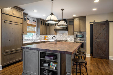 Inspiration for a large transitional l-shaped medium tone wood floor eat-in kitchen remodel in Chicago with a drop-in sink, recessed-panel cabinets, gray cabinets, wood countertops, white backsplash, ceramic backsplash, paneled appliances and an island
