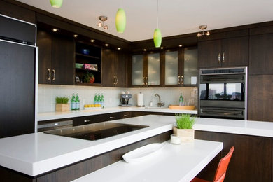 Example of a mid-sized trendy l-shaped bamboo floor and brown floor kitchen design in Milwaukee with an undermount sink, flat-panel cabinets, dark wood cabinets, solid surface countertops, white backsplash, black appliances, an island and glass tile backsplash