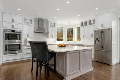 This is an example of a large traditional u-shaped kitchen/diner in Bridgeport with a belfast sink, shaker cabinets, white cabinets, engineered stone countertops, multi-coloured splashback, marble splashback, stainless steel appliances, dark hardwood flooring, an island, brown floors and white worktops.
