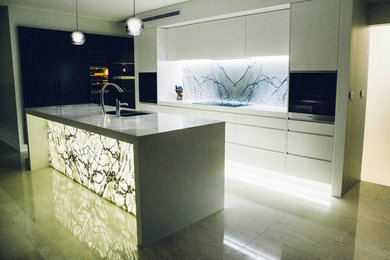 Modern inredning av ett linjärt kök och matrum, med vita skåp, vitt stänkskydd, stänkskydd i sten, en köksö, en undermonterad diskho, släta luckor, marmorbänkskiva och klinkergolv i porslin