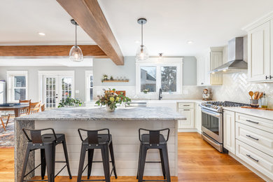 Photo of a medium sized classic l-shaped kitchen/diner in Boston with a belfast sink, raised-panel cabinets, white cabinets, engineered stone countertops, white splashback, metro tiled splashback, stainless steel appliances, light hardwood flooring, an island, beige floors and white worktops.