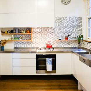 Small contemporary l-shaped kitchen in Newcastle - Maitland with an integrated sink, flat-panel cabinets, white cabinets, stainless steel benchtops, white splashback, mosaic tile splashback, stainless steel appliances, medium hardwood floors, brown floor and grey benchtop.