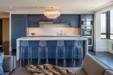 Traditional galley open plan kitchen in San Francisco with shaker cabinets, blue cabinets, white splashback, stone slab splashback, integrated appliances, dark hardwood flooring and an island.