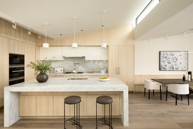 Bild på ett funkis vit vitt l-kök, med en undermonterad diskho, släta luckor, skåp i ljust trä, grått stänkskydd, stänkskydd i sten, rostfria vitvaror, mellanmörkt trägolv, en köksö och brunt golv