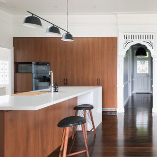 This is an example of a contemporary l-shaped kitchen in Brisbane with flat-panel cabinets, medium wood cabinets, black appliances, medium hardwood floors, with island, brown floor and white benchtop.