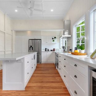 Design ideas for a country l-shaped kitchen in Brisbane with shaker cabinets, white cabinets, white splashback, window splashback, stainless steel appliances, medium hardwood floors, with island, brown floor and white benchtop.