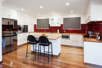 Modelo de cocina actual con encimera de madera, salpicadero rojo, salpicadero de vidrio templado, electrodomésticos de acero inoxidable y suelo de madera clara