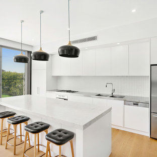 This is an example of a mid-sized contemporary single-wall open plan kitchen in Sydney with an integrated sink, flat-panel cabinets, white cabinets, stainless steel benchtops, white splashback, stainless steel appliances, medium hardwood floors, with island and brown floor.