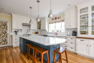 Example of a mid-sized transitional u-shaped light wood floor eat-in kitchen design in Portland Maine with a farmhouse sink, shaker cabinets, white cabinets, solid surface countertops, white backsplash, subway tile backsplash, stainless steel appliances and an island