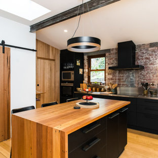 Inspiration for an industrial kitchen in Hobart with flat-panel cabinets, black cabinets, stainless steel benchtops, brick splashback, light hardwood floors, with island and beige floor.