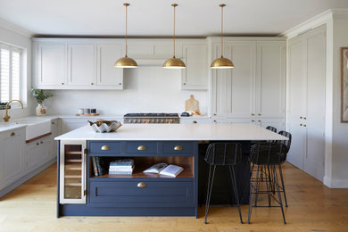 Photo of a traditional l-shaped kitchen in London with a belfast sink, shaker cabinets, grey cabinets, white splashback, stainless steel appliances, medium hardwood flooring, an island, brown floors and white worktops.