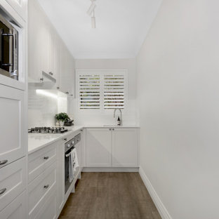 This is an example of a transitional l-shaped kitchen in Sydney with an undermount sink, shaker cabinets, white cabinets, white splashback, glass tile splashback, stainless steel appliances, light hardwood floors, no island, brown floor and white benchtop.