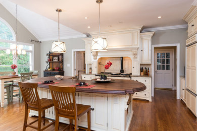 Large transitional l-shaped medium tone wood floor and brown floor eat-in kitchen photo in Raleigh with a farmhouse sink, raised-panel cabinets, beige cabinets, wood countertops, beige backsplash, porcelain backsplash, paneled appliances, an island and brown countertops