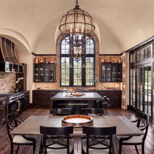 Diseño de cocina comedor en L, clásica, grande, con salpicadero beige, salpicadero de ladrillos, suelo de madera oscura, armarios con paneles empotrados, puertas de armario de madera en tonos medios, electrodomésticos negros y una isla