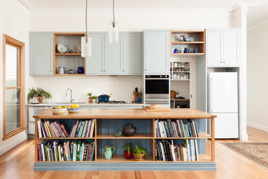 Foto på ett mellanstort kök, med en dubbel diskho, skåp i shakerstil, blå skåp, träbänkskiva, vitt stänkskydd, stänkskydd i tunnelbanekakel, rostfria vitvaror, ljust trägolv och en köksö
