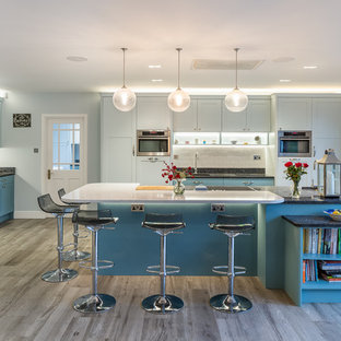 Design ideas for a large classic u-shaped kitchen in Hertfordshire with a built-in sink, shaker cabinets, blue cabinets, granite worktops, white splashback, stone slab splashback, stainless steel appliances, medium hardwood flooring, black worktops, an island and grey floors.