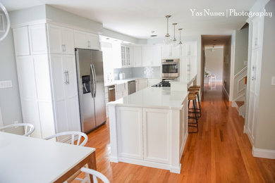 Large transitional u-shaped medium tone wood floor eat-in kitchen photo in Boston with a farmhouse sink, flat-panel cabinets, white cabinets, quartz countertops, white backsplash, glass tile backsplash, stainless steel appliances and an island