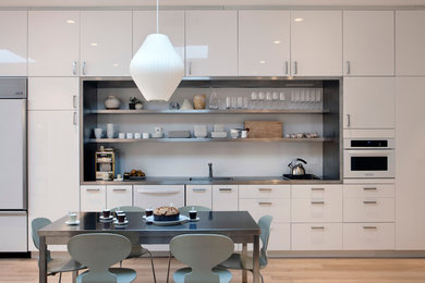 Trendy single-wall eat-in kitchen photo in New York with white appliances, flat-panel cabinets, stainless steel countertops, white cabinets and an integrated sink