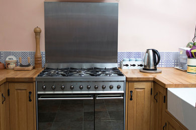 This is an example of a small rustic u-shaped kitchen/diner in Surrey with a belfast sink, raised-panel cabinets, medium wood cabinets, wood worktops, multi-coloured splashback, porcelain splashback, stainless steel appliances, grey floors and brown worktops.