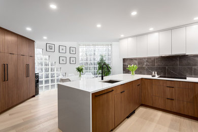 Bild på ett mellanstort funkis vit linjärt vitt kök och matrum, med släta luckor, svarta vitvaror, ljust trägolv, en halv köksö, en undermonterad diskho, skåp i ljust trä, bänkskiva i kvartsit, svart stänkskydd, stänkskydd i stenkakel och brunt golv