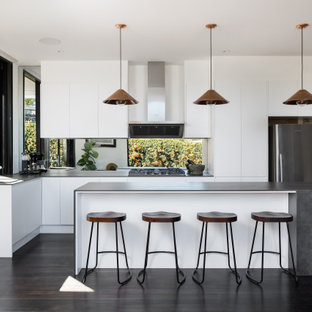 Contemporary galley open plan kitchen in Sydney with a drop-in sink, white cabinets, solid surface benchtops, mirror splashback, stainless steel appliances, painted wood floors, with island, black floor and grey benchtop.