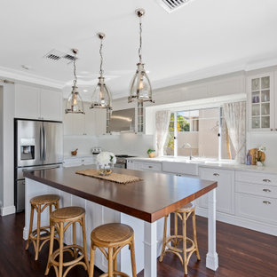 This is an example of a large transitional l-shaped eat-in kitchen in Gold Coast - Tweed with a farmhouse sink, shaker cabinets, white cabinets, wood benchtops, white splashback, porcelain splashback, stainless steel appliances, dark hardwood floors, with island, brown floor and brown benchtop.