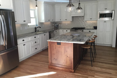 Example of a large classic l-shaped medium tone wood floor and brown floor open concept kitchen design in Other with raised-panel cabinets, white cabinets, granite countertops, an island, an undermount sink, white backsplash, subway tile backsplash, stainless steel appliances and blue countertops