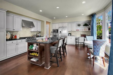 Example of a mid-sized classic dark wood floor and brown floor eat-in kitchen design in Seattle with an undermount sink, raised-panel cabinets, white cabinets, quartz countertops, gray backsplash, stone tile backsplash, stainless steel appliances and an island