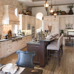 Imagen de cocina lineal, tradicional, grande, cerrada, con electrodomésticos con paneles, fregadero sobremueble, armarios con paneles con relieve, puertas de armario con efecto envejecido, encimera de granito, salpicadero marrón, salpicadero de travertino, suelo de madera en tonos medios, una isla y suelo marrón