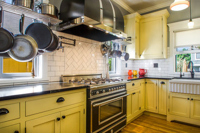 Design ideas for a medium sized traditional kitchen in Orange County with a belfast sink, shaker cabinets, yellow cabinets, soapstone worktops, white splashback, ceramic splashback, black appliances and medium hardwood flooring.