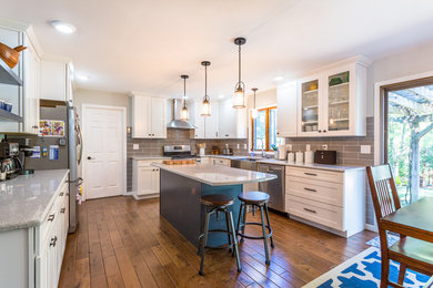 Example of a transitional u-shaped medium tone wood floor eat-in kitchen design in Indianapolis with a farmhouse sink, shaker cabinets, white cabinets, gray backsplash, subway tile backsplash, stainless steel appliances and an island