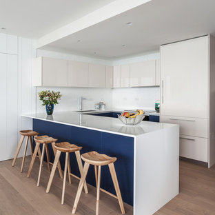 Design ideas for a beach style u-shaped kitchen in Miami with an undermount sink, flat-panel cabinets, beige cabinets, white splashback, panelled appliances, light hardwood floors, a peninsula and white benchtop.