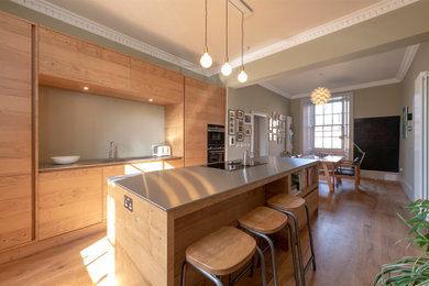 Medium sized contemporary galley kitchen/diner in Edinburgh with flat-panel cabinets, medium wood cabinets, stainless steel worktops, grey splashback, black appliances and an island.