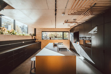 This is an example of a large modern kitchen in Other with a built-in sink, flat-panel cabinets, black cabinets, stainless steel worktops, black splashback, wood splashback, black appliances, concrete flooring, an island, grey floors and grey worktops.