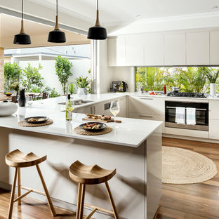 Design ideas for a contemporary u-shaped open plan kitchen in Perth with a double-bowl sink, flat-panel cabinets, beige cabinets, window splashback, stainless steel appliances, medium hardwood floors, a peninsula and brown floor.